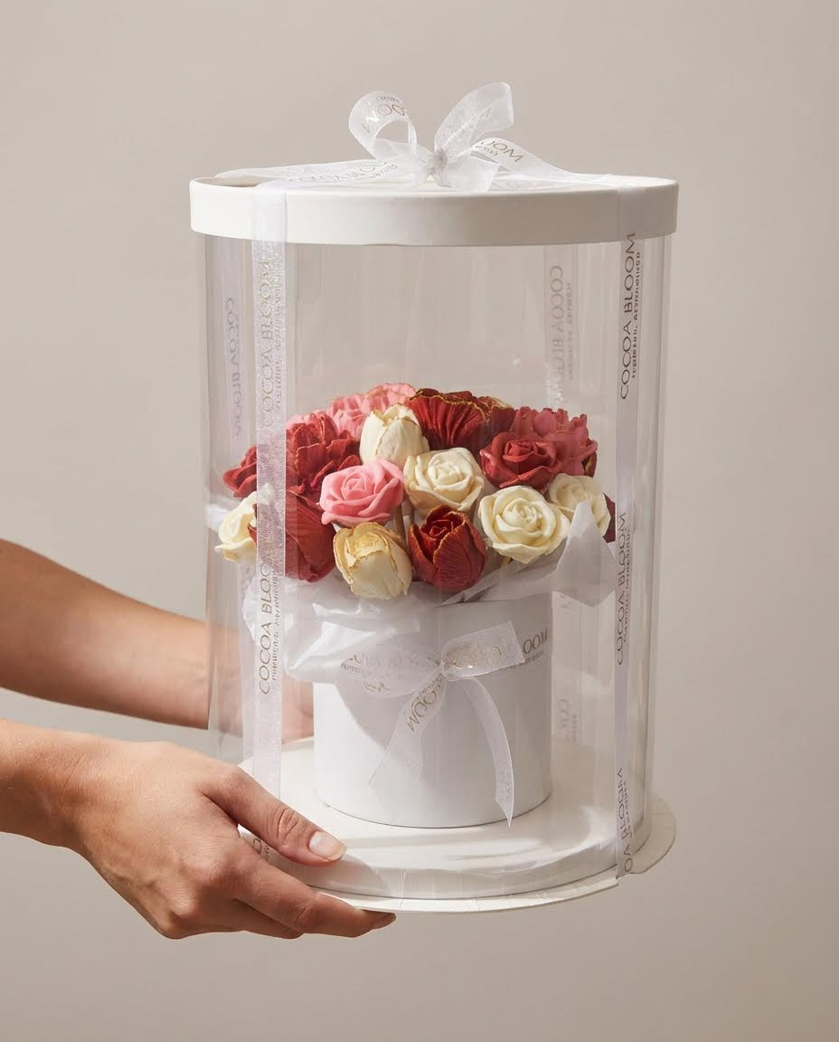 Hand holding a cylindrical gift box with a bouquet of red, pink, and white 3D chocolate roses on a neutral background